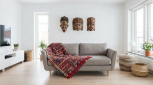 Modern Scandinavian living room featuring a grey sofa with a colorful Moroccan kilim rug draped over it. Above the sofa is a gallery wall of three carved wooden African and Asian masks. The room has light wood floors and a large window.